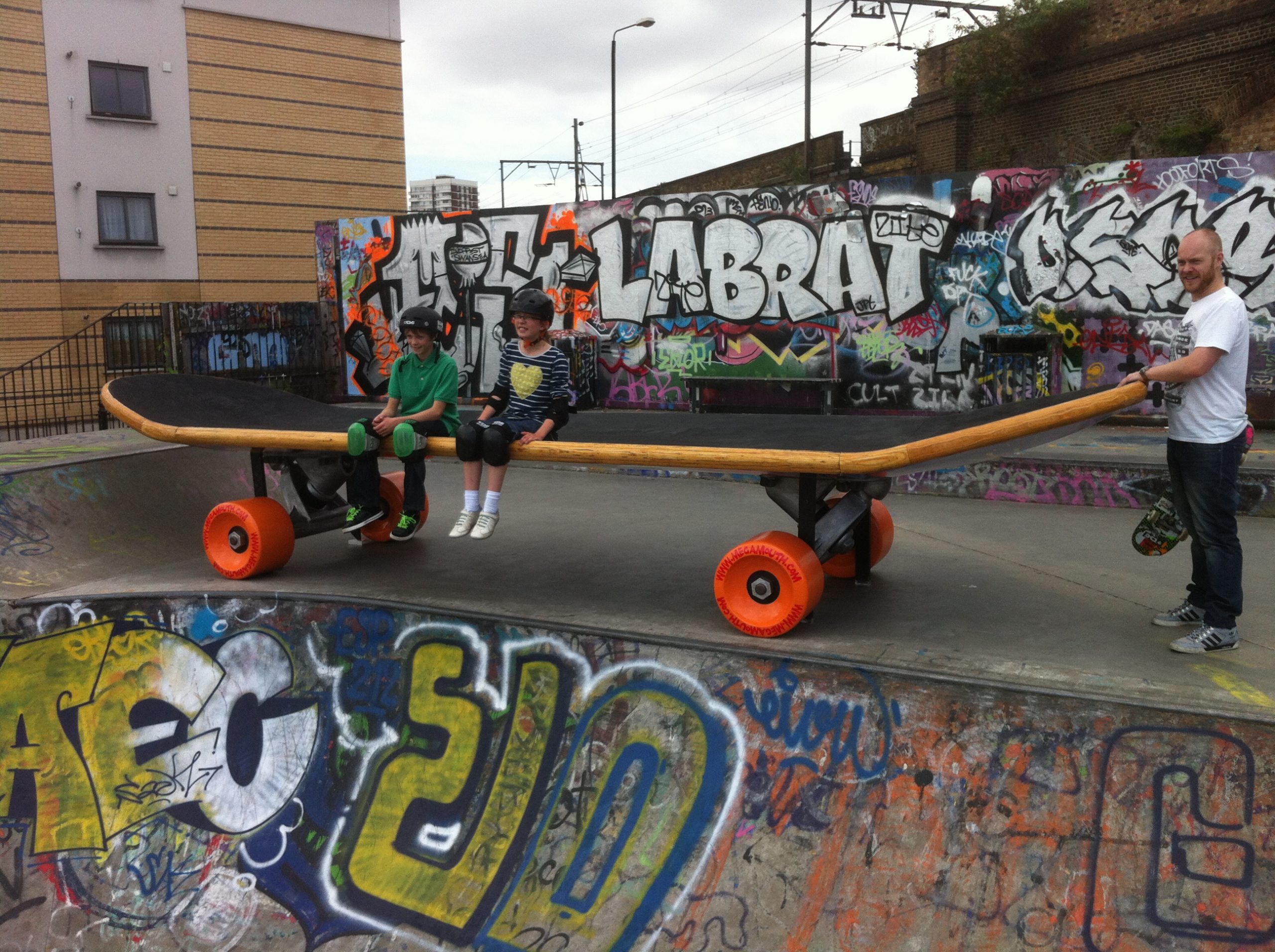 A model of a large skateboard in a skate park with people sat on top